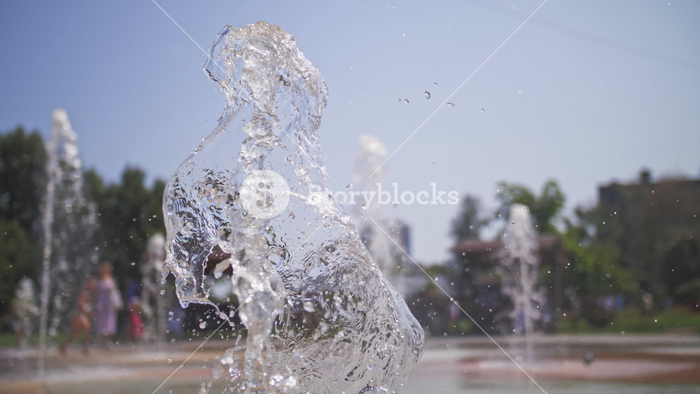 Refreshing water sprays of city fountain against background of having fun children. Escape from heat in park during summer vacation.