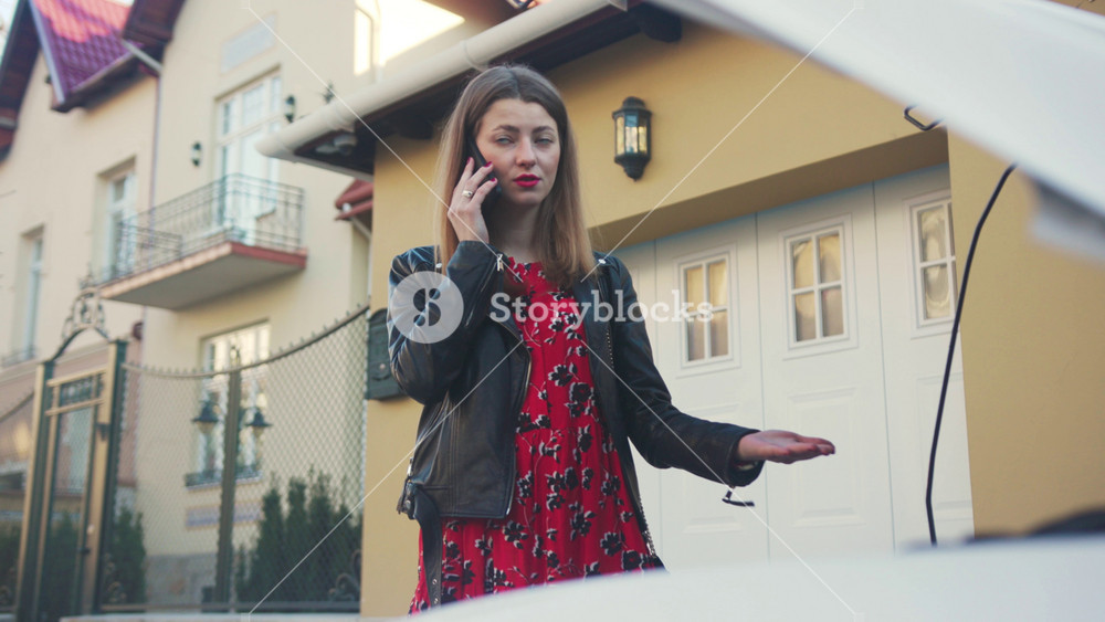 Graceful young Caucasian woman wearing standing near broken car and calling friend for help. Portrait of classy unhappy girl. Yellow house