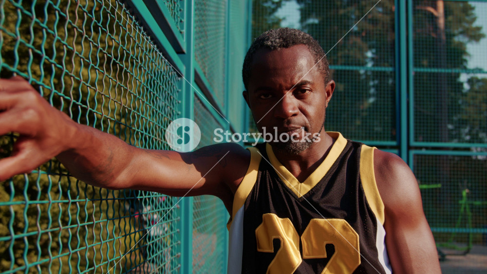 Portrait of serious tired Afro American basketball player with beard in stylish sportswear standing in basketball court looking directly at
