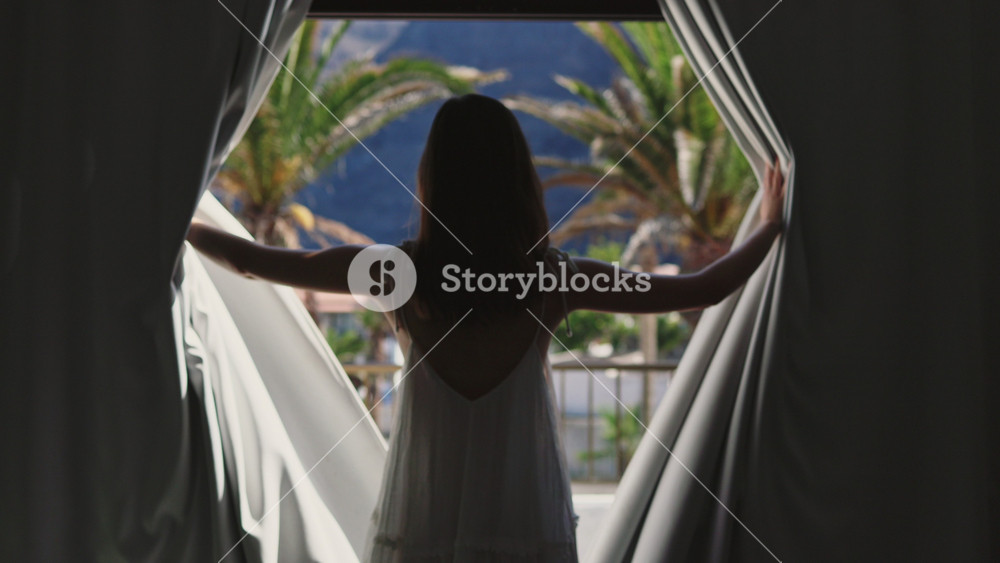 In dark room young woman opening window curtains inside apartment. Pretty tender girl in long beautiful white dress walking on terrace