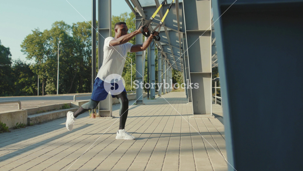 NEW YORK - September 5, 2020: Sporty african american young guy working out with trx fitness rope outdoors body exercises at stadium