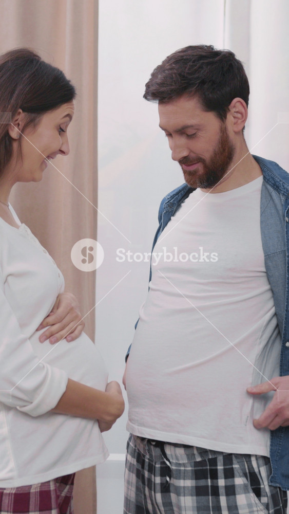 Lovely young woman expecting child laughing at her pregnant husband. Pretending funny man showing food tummy. Families fun.
