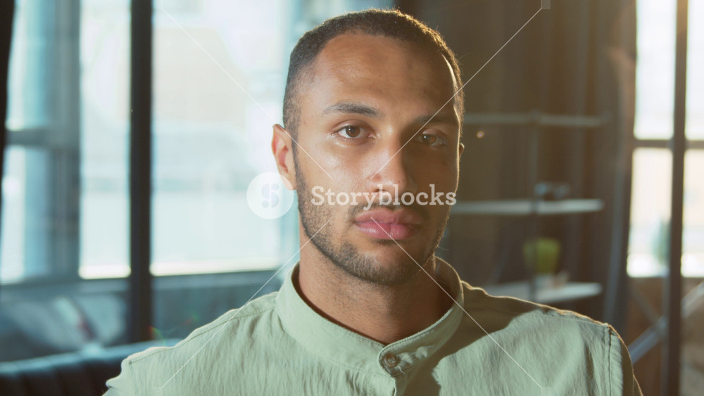 Close-up portrait of serious young African American unshaven man with brown eyes looking directly at camera on blurred background of modern