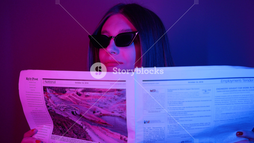 NEW YORK - April 5, 2019: Shooting of enchanting young woman in black sunglasses holding newspaper in hands, looking around. Attractive spy