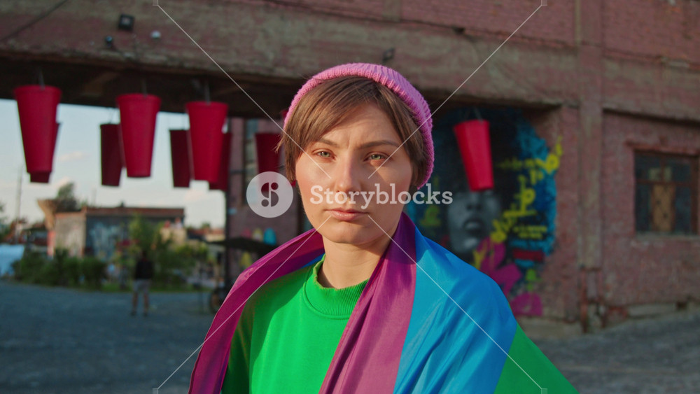 Horizontal shot of serious fashionable lesbian woman wearing rainbow flag, standing in street with old building. Hipster girl in colorful