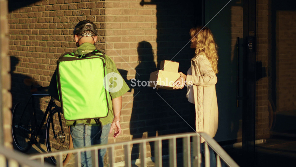 Slow motion man courier in a safety helmet delivering food in a paper bag to the customer home by bicycle at sunlight attractive woman stand