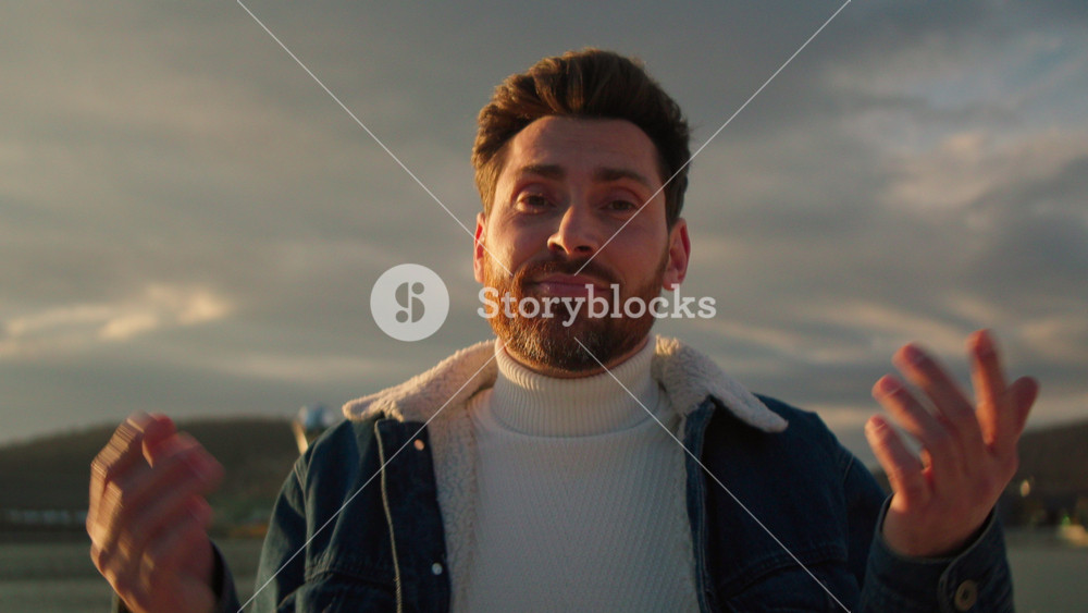 Portrait of good-looking young bearded man in casual clothes looking at camera and shrugging on background of lake with beautiful sunset