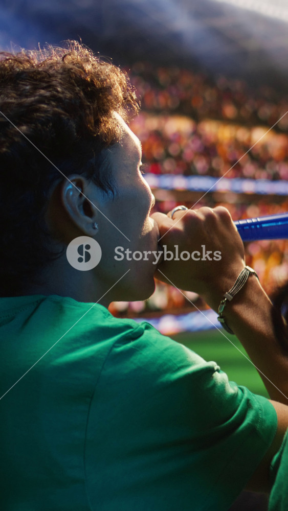 Vertical video Close up of fans using vuvuzela in stadium tribunes during football match. Back view of supporter blowing air next to woman