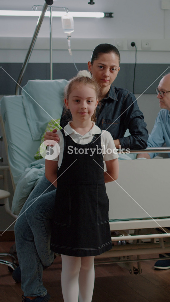 Vertical video: Portrait of child and mother visiting old patient in hospital ward bed. Woman and kid sitting in intensive care room at