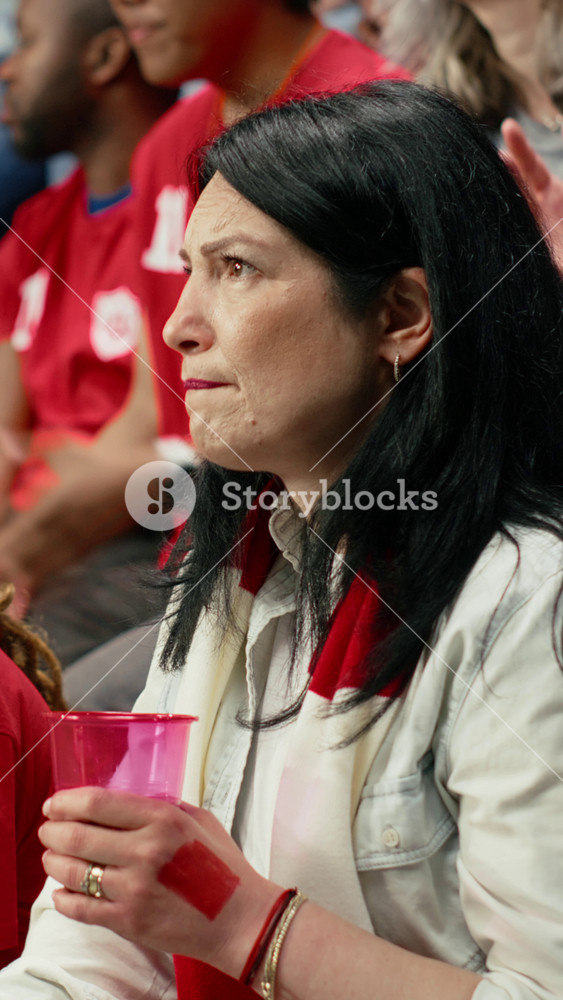 Vertical Video Worried mature female football supporter holding pink cup and red scarf in stadium stands during tense live soccer match