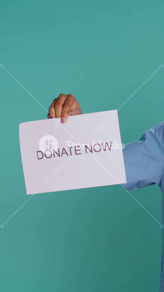 Vertical video Portrait of male nurse holding donate now sign, inviting people to contribute, studio background. Healthcare worker in need