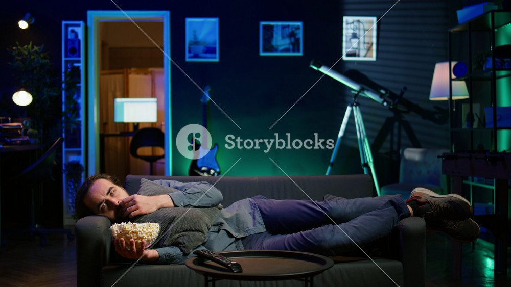 Depressed man at home lounged on couch feeling low, seeking comfort by watching movies on TV and eating popcorn. Glum person laying down