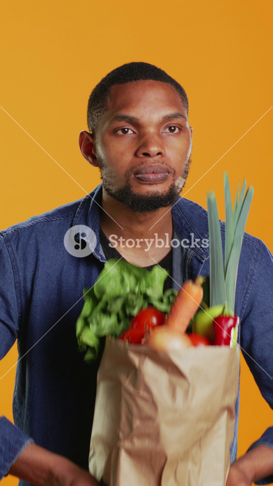 Vertical Video Young adult pretending to lift weight with a paper bag full of bio groceries, mimicking workout exercises in studio. Male