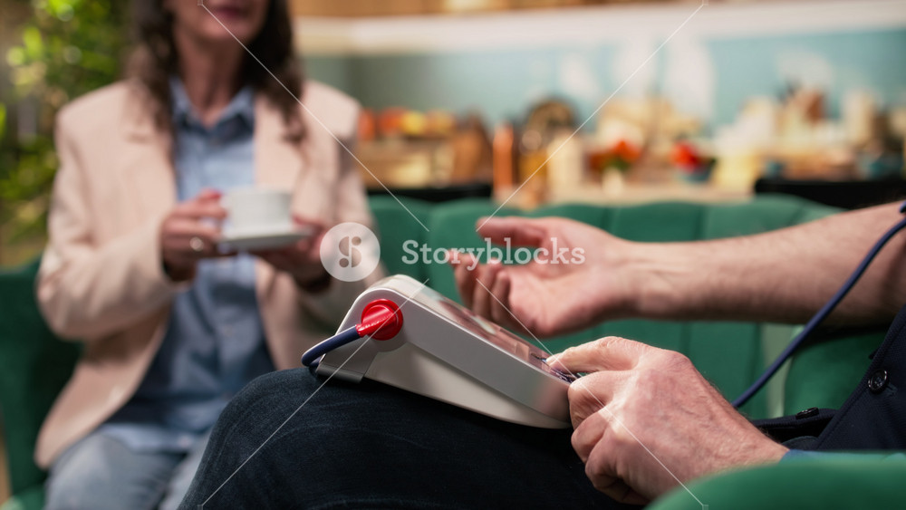 Senior man checking his blood pressure on monitor, while his wife drinking a cup of drink in their living room at home.