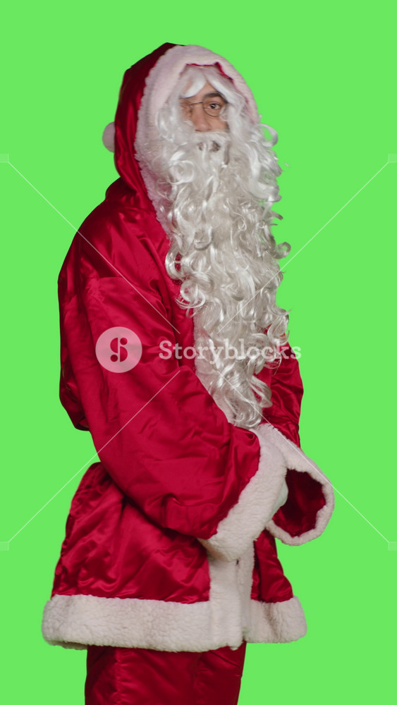 Vertical video Profile of adult in costume looks at wristwatch to check time against greenscreen backdrop, wearing santa claus red festive