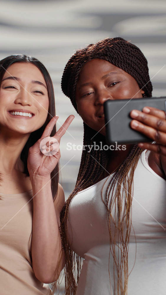 Vertical video: Happy flawless girls taking photos on mobile phone, using smartphone to have fun with pictures. Curvy and skinny ladies