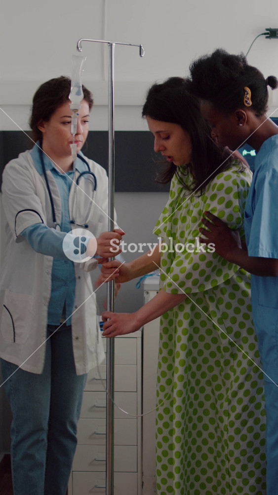 Vertical video: Medical team helping patient to stand up from bed holding intravenous IV fluid drip bag after recovery surgery in hospital