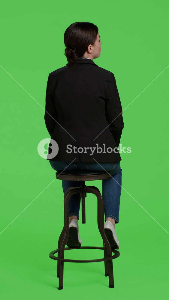Vertical video: Back view of company employee sitting on chair and waiting on camera, full body greenscreen backdrop. Young businesswoman in