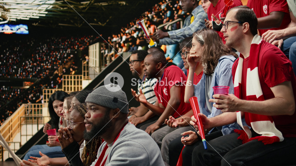 Diverse red team soccer fans watching live football match from stadium stands, seated supporters reacting with focus and team spirit