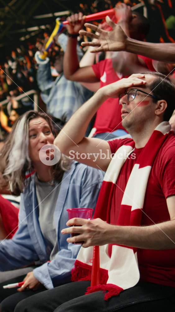 Vertical Video Red team soccer fans reacting to close call in stadium stands, hands on head, scarves and anxious crowd during live football