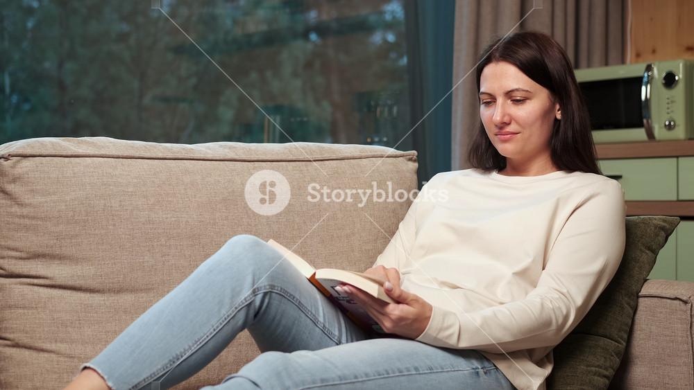 Brunette woman sitting comfortably on living room couch, reading captivating book near sunlit window, enjoying peaceful moment of quiet