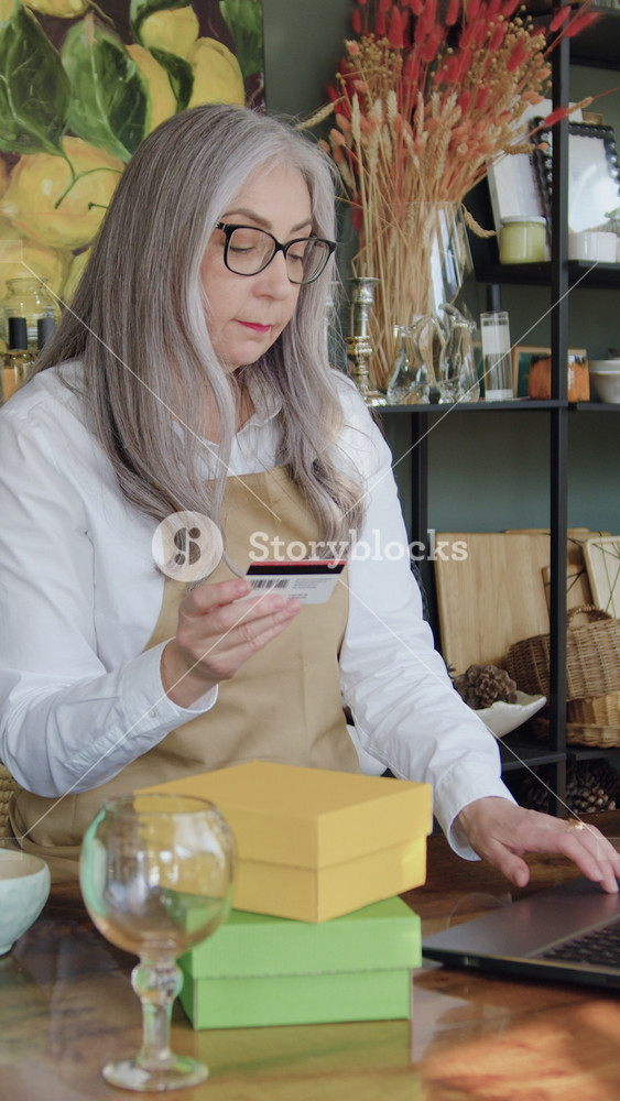 Woman with long straight grey hair in glasses sitting at her workplace in contemporary small gift shop and writing bank account datas for