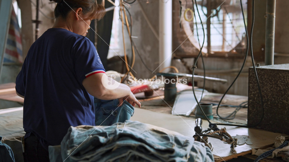 China, Guangzhou 01 August 2026 Factory Female Worker Irons New Denim Jeans with Heavy Industrial Steam Iron. One Garment Finishing Stage in