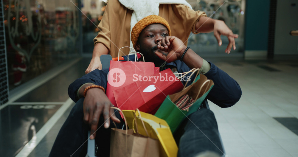 Happy afro-american excited guy riding in shopping cart with girlfriend showing places to buy gifts. Carefree lifestyle. Fun concept. Crazy