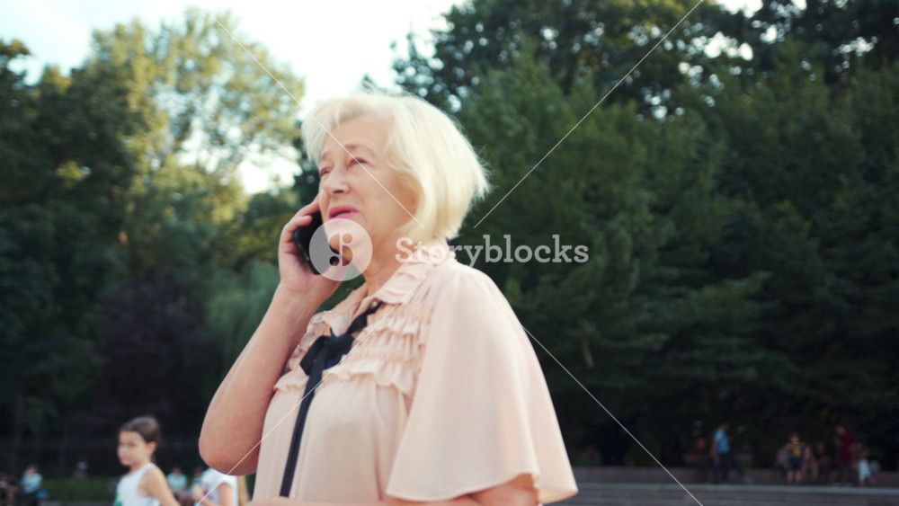 Elegant Caucasian young woman standing near park and talking on cell phone. Healthy pensioner with blonde hair smiling, Conversation