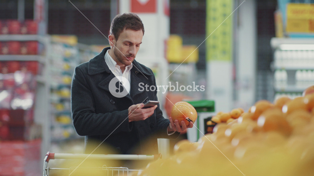Smiling young man walking in supermarket with shopping trolley which looking on shopping list in phone choose fruit buyer customer business