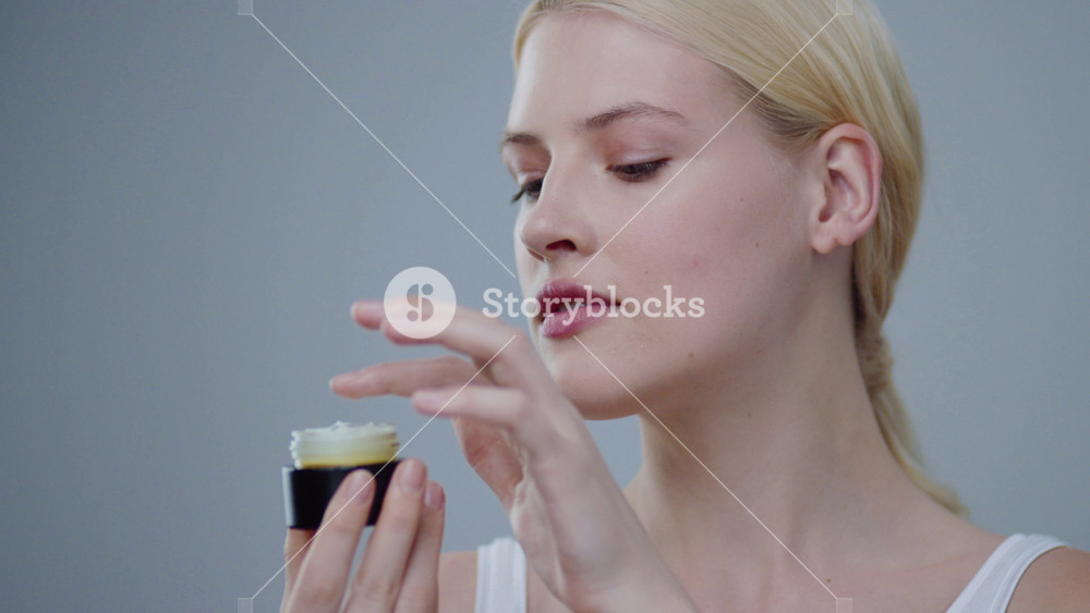 Close-up of stunning blonde young woman with perfect skin putting on moisturizing cream and smiling on camera against grey background. Skin