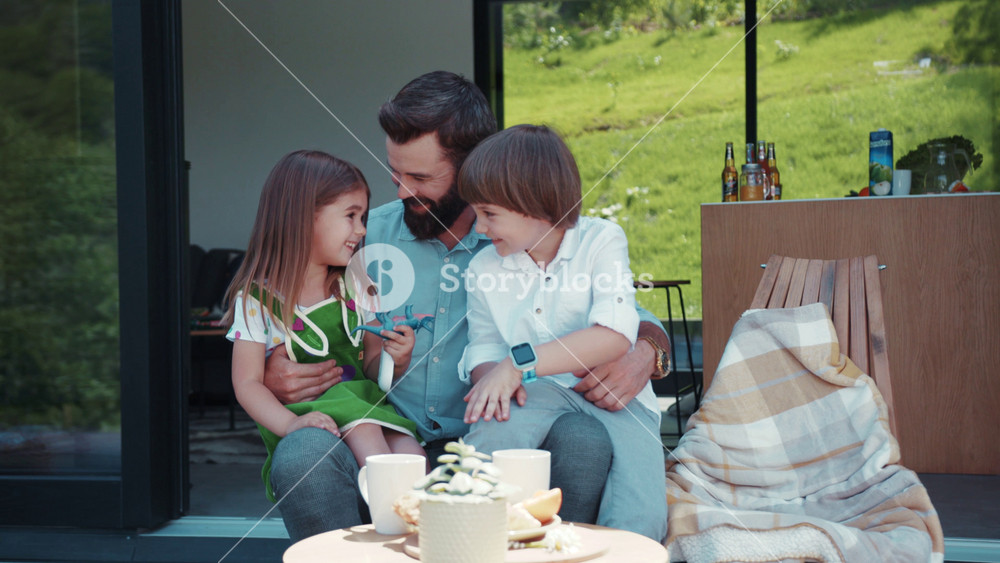 Little joyful cute boy and girl sit on father’s knees, talking, laughing at his jokes. Happy childhood, time for family, family bonds