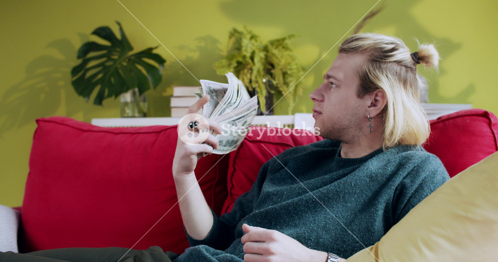 Satisfied young blonde businessman celebrating payday holding bunch of money waving like fan relaxing on couch in living room. At home