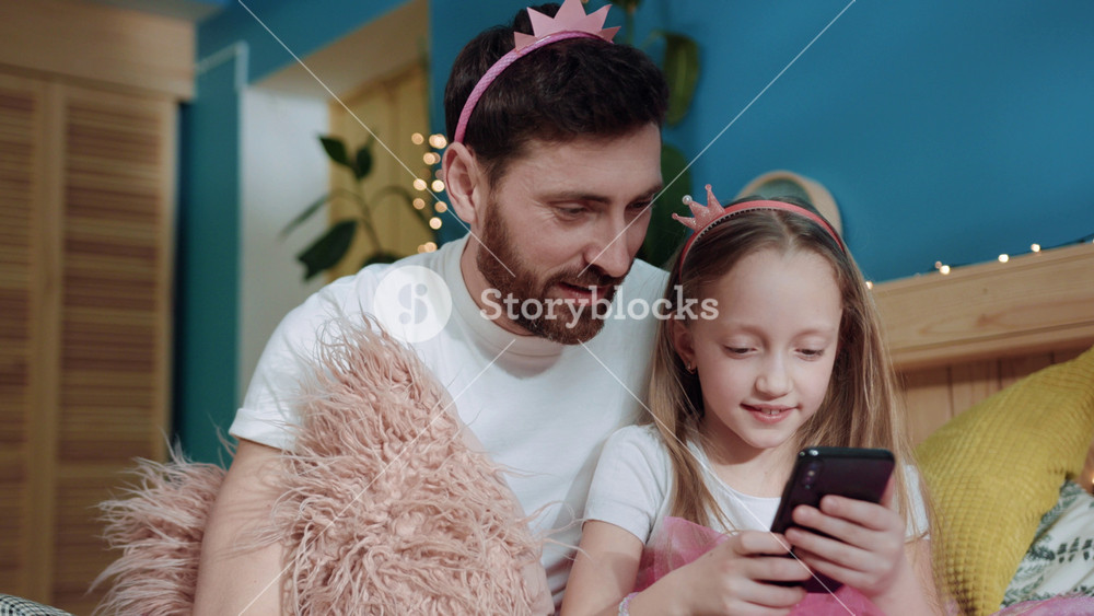 Closeup portrait excited cute girl leaning on her father dressed like fairies watching cartoons together om smartphone sitting on bed at