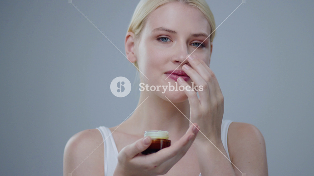 Joyful blonde woman holding a cream jar puts a drop on her nose and laughs on camera. Portrait of amazing female model with angel skin