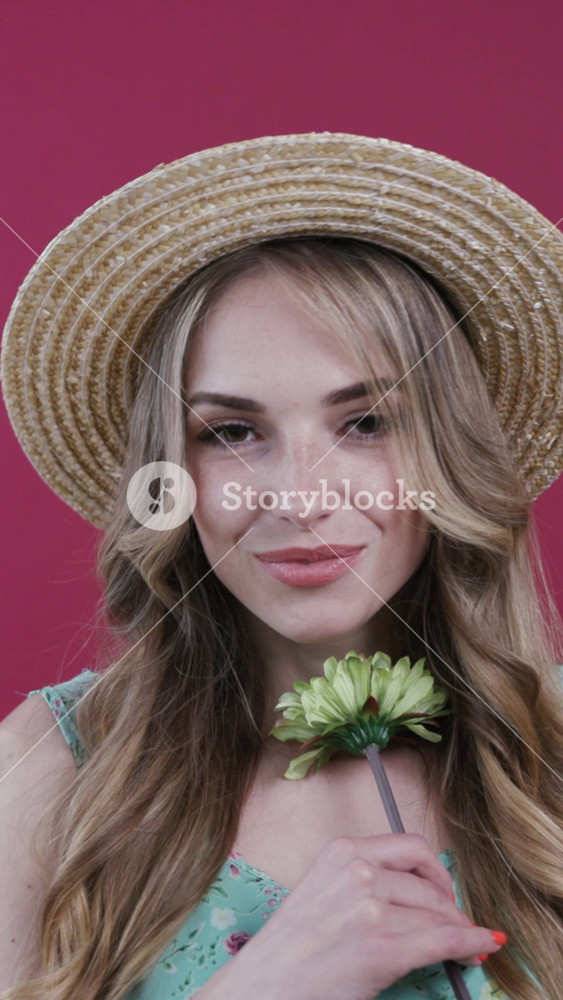 Close up view of good-looking blonde woman with wavy hair in a trendy summer straw hat smelling the flower, turning to camera and lovely