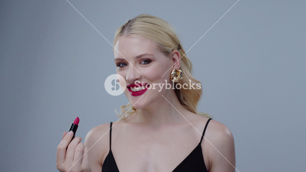 Stunning blonde model girl with red lips applying lipstick looking in the mirror. Portrait of attractive young woman smiling broadly