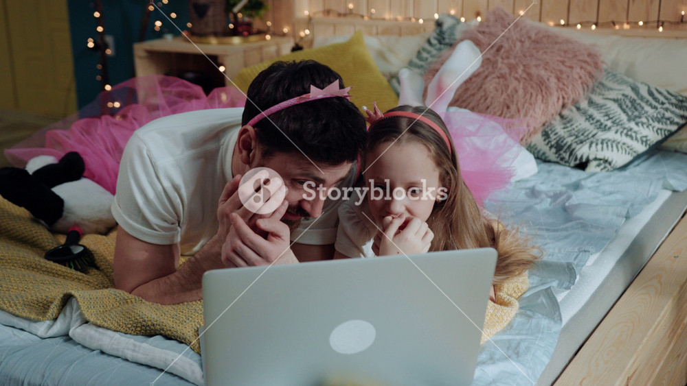 Closeup family portrait young dad and girl dressed like fairies watching scary cartoons together on laptop computer lying on bed at home