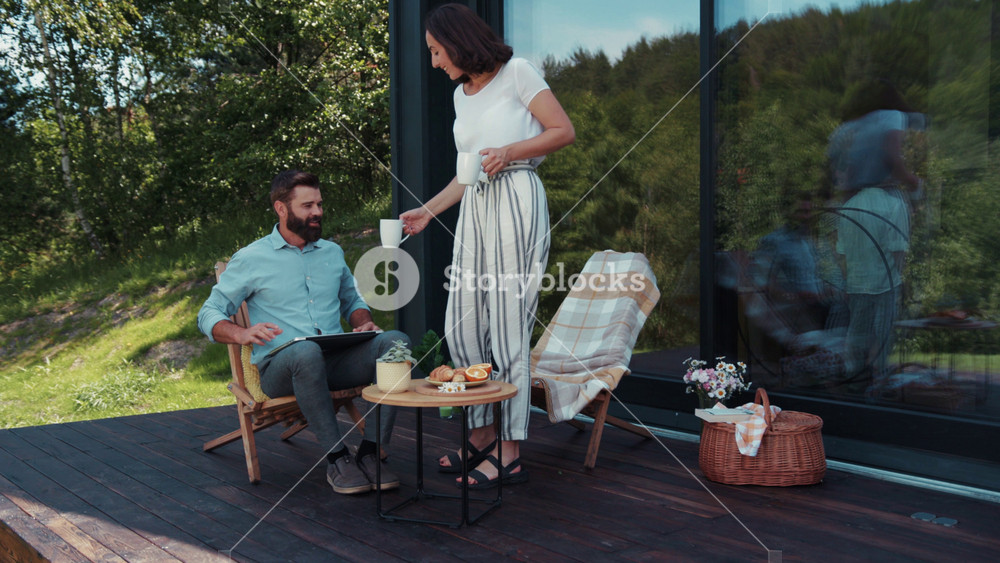 Beautiful caring young woman in trendy outfit serves coffee to a bearded man who actively uses laptop on the cozy terrace. Happy, family