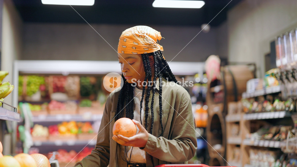 Concentrated afro-american customer young woman walking in natural fruit section of supermarket choosing fresh oranges for food shopping.