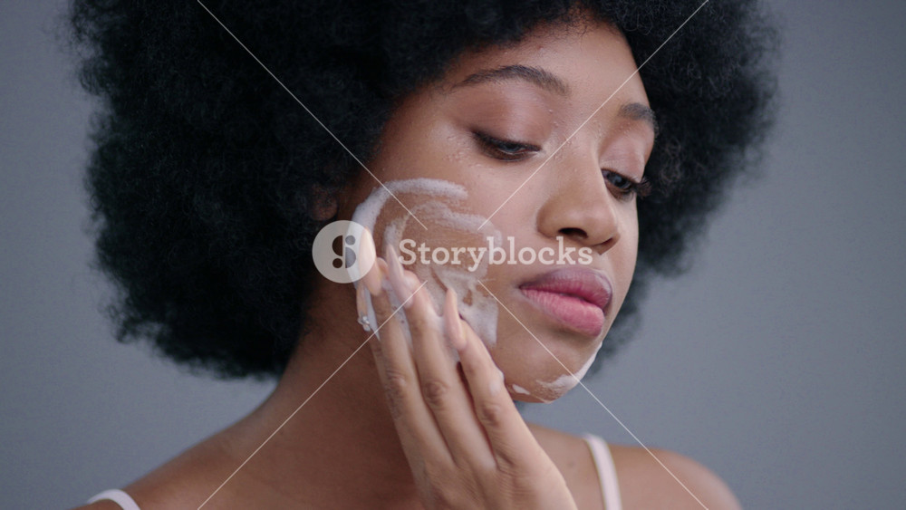 Portrait of amazing afro girl applying moisturizing foam on her clear face on grey background. Close-up of attractive young woman model