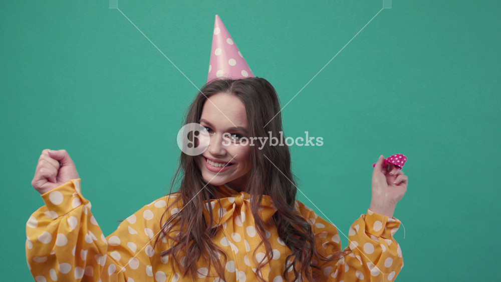 Attractive curly woman in a birthday hat uses birthday horn, cheers up on a green background. Having fun, birthday party, cheerful mood