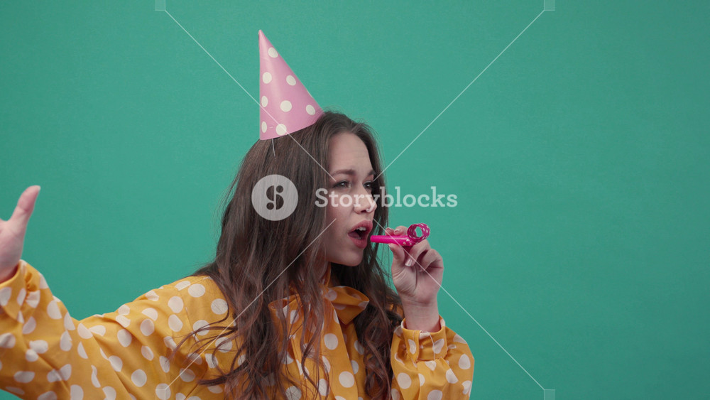 Cute young woman in a birthday hat hilariously uses birthday horn, cheers up on a green background. Having party, cheerful mood. Slow motion
