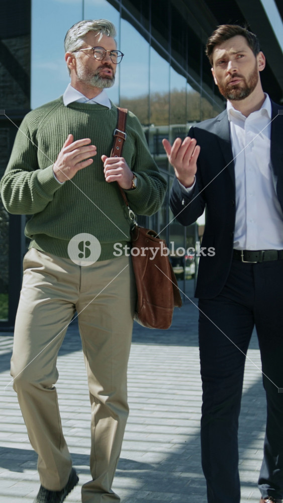 Two good-looking male colleagues walking near business center, talking and gesturing. Front view of Caucasian men in elegant business