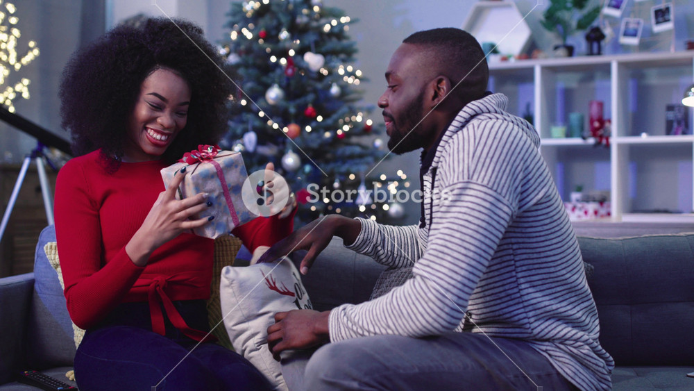 Nice African couple of young people talking and smiling. Boyfriend giving holiday gift to girl. Grateful girl hugging guy. Indoors. Evening.