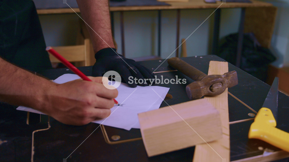 Carpenter wearing black gloves drawing sketch of furniture with red pencil on sheet of paper placed on workbench, near wooden block, hammer