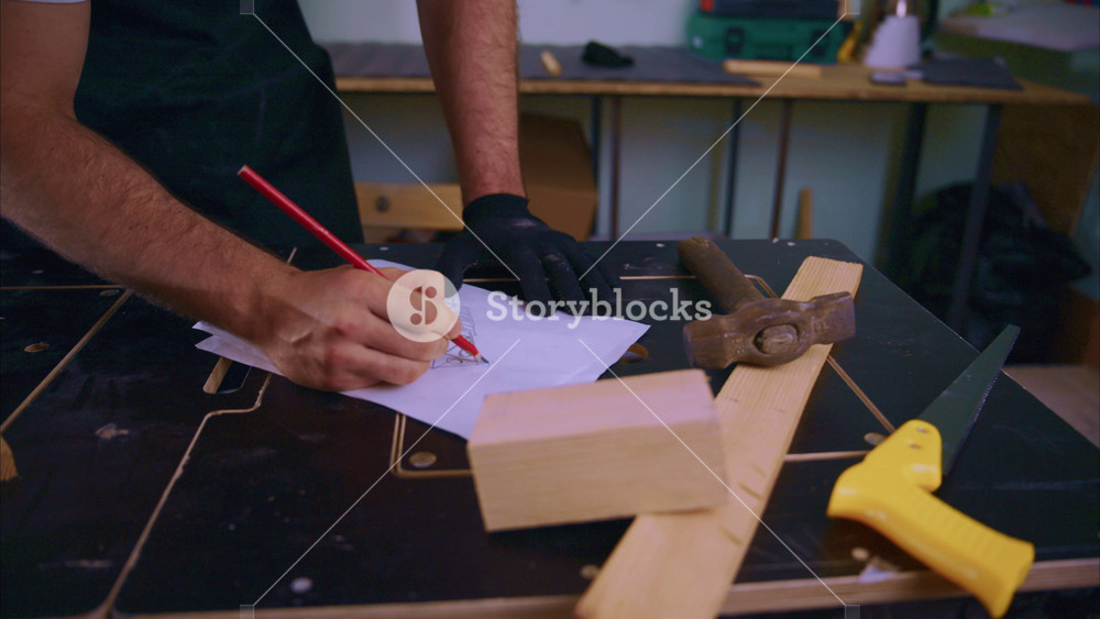 Carpenter wearing black gloves drawing sketch of furniture with red pencil on sheet of paper placed on workbench with wooden block, plank