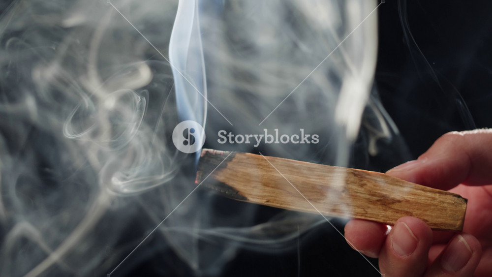 Person holding Palo Santo wood stick in hand, aromatic Palo Santo burning on black background, smoke spreading around, slow motion video
