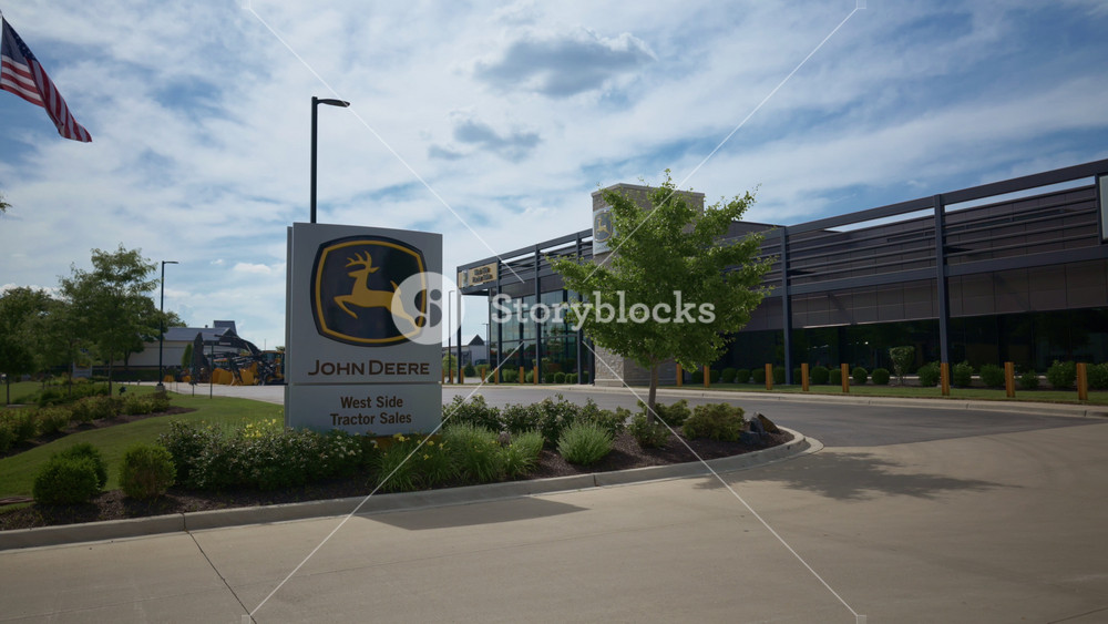 The exterior of John Deere retail center with sunny blue sky. The dealership building displays the John Deere logo and branding. - June 27