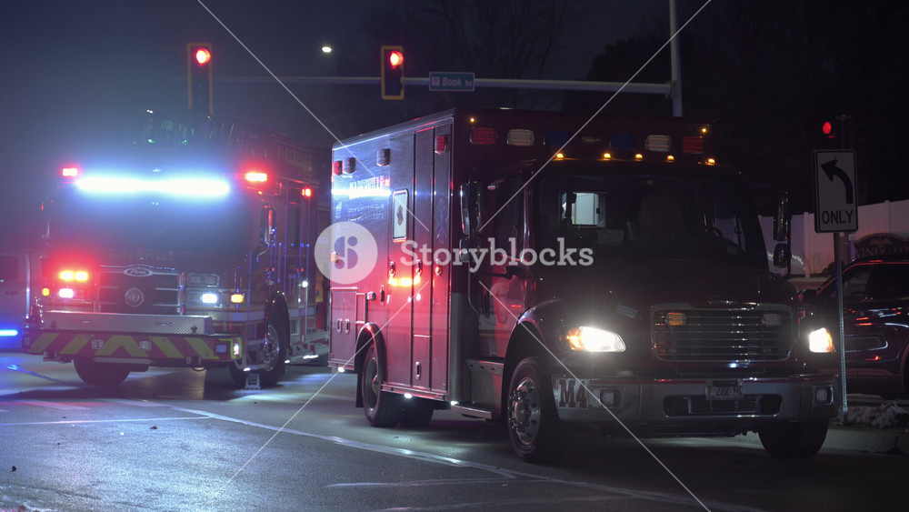 POV Nighttime scene of emergency vehicles working at a roadside incident. Police cars, fire trucks, ambulances, cars stopping at a red light